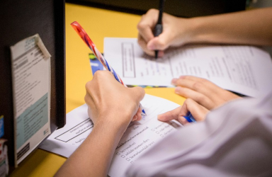 A person writing on paper with pens and pencils.